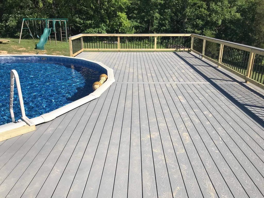 Curved backyard pool with blue water and gray deck beside a wooden railing and green trees