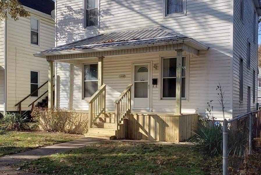 Front porch of a beige house with steps, railing, and a small front yard in sunlight