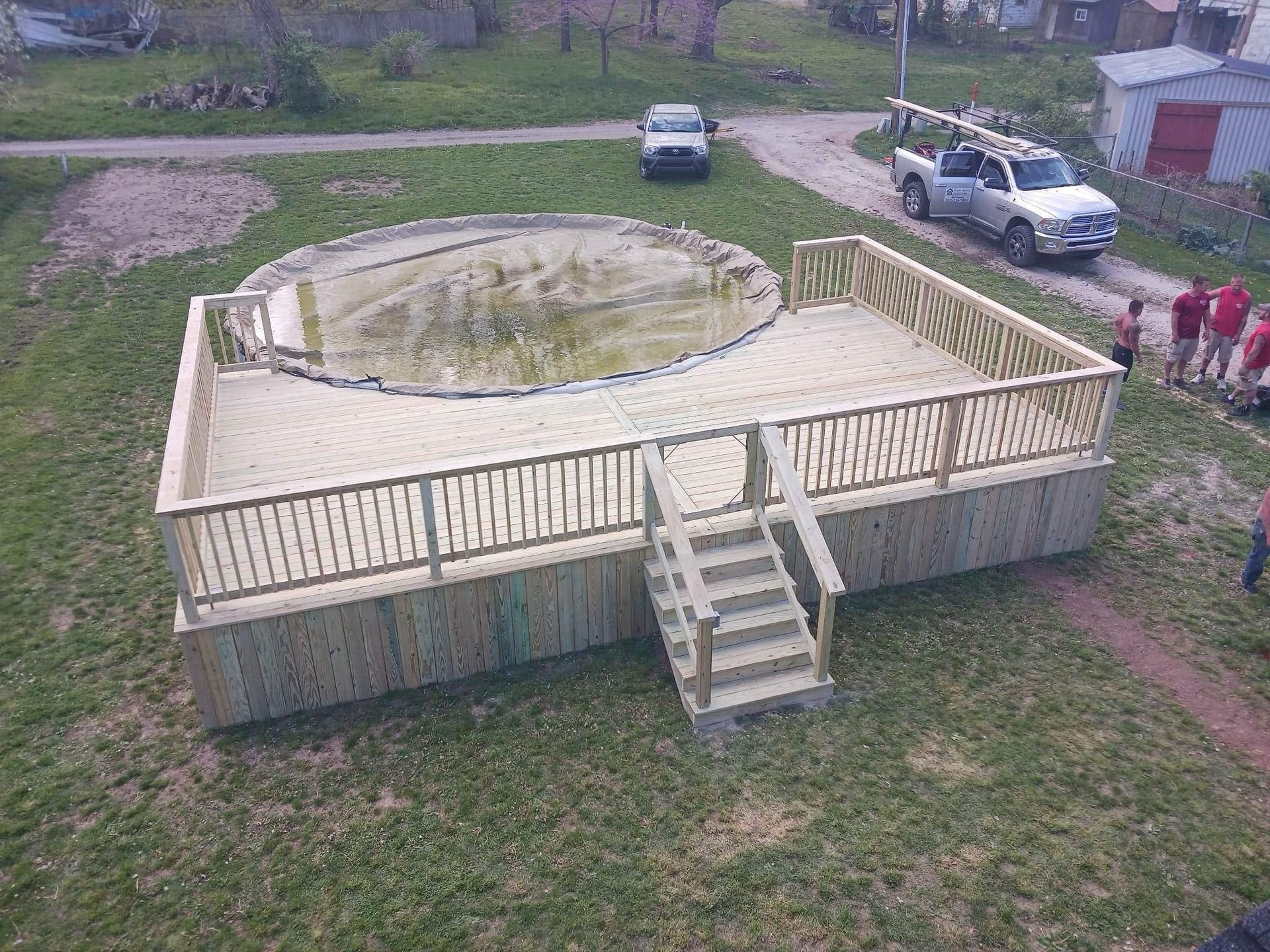 Small wooden deck surrounding an above-ground pool, with stairs and a few parked vehicles nearby.