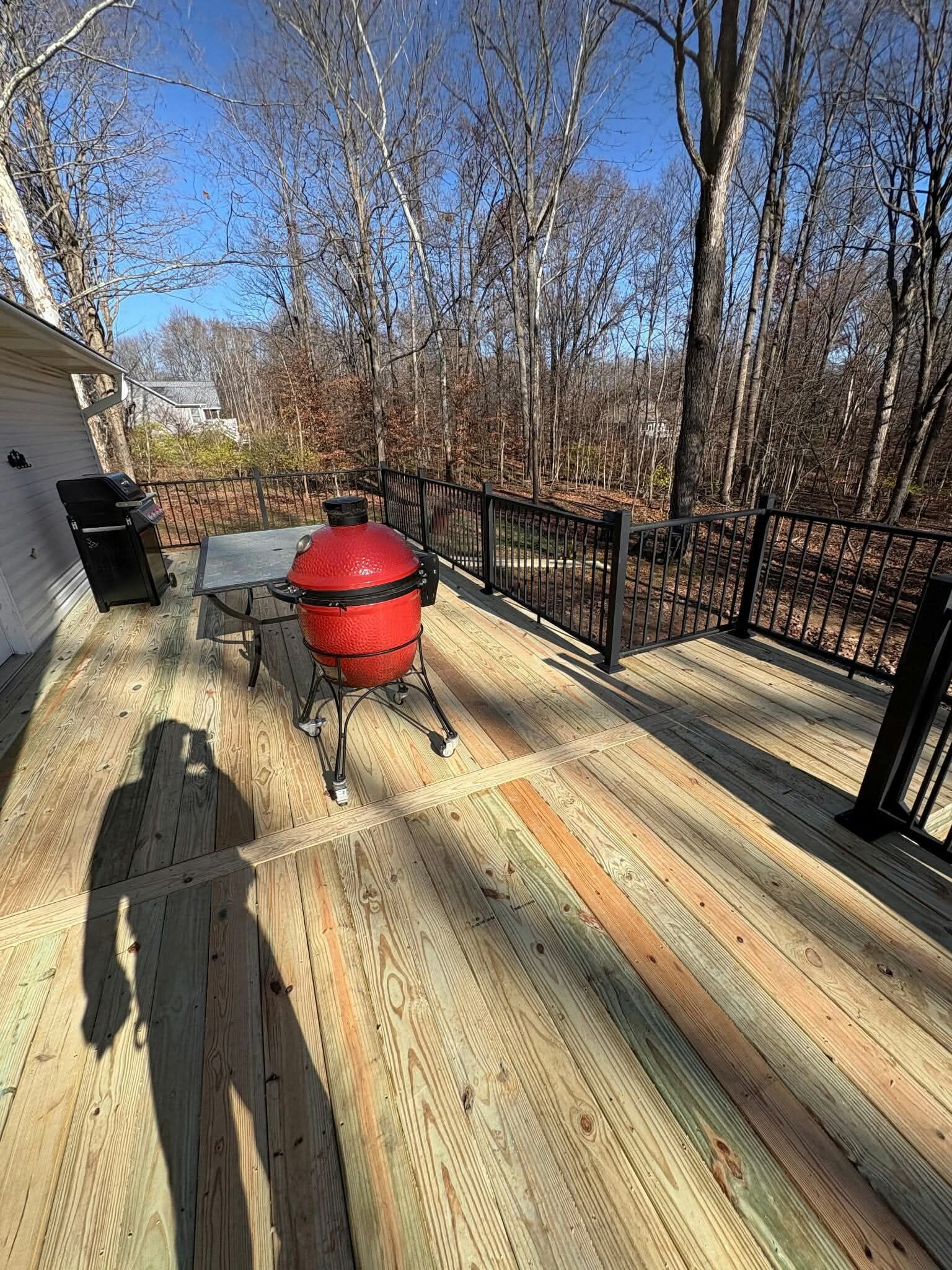 Red outdoor smoker on a wooden deck beside a black railing and leafless trees under a bright blue sky