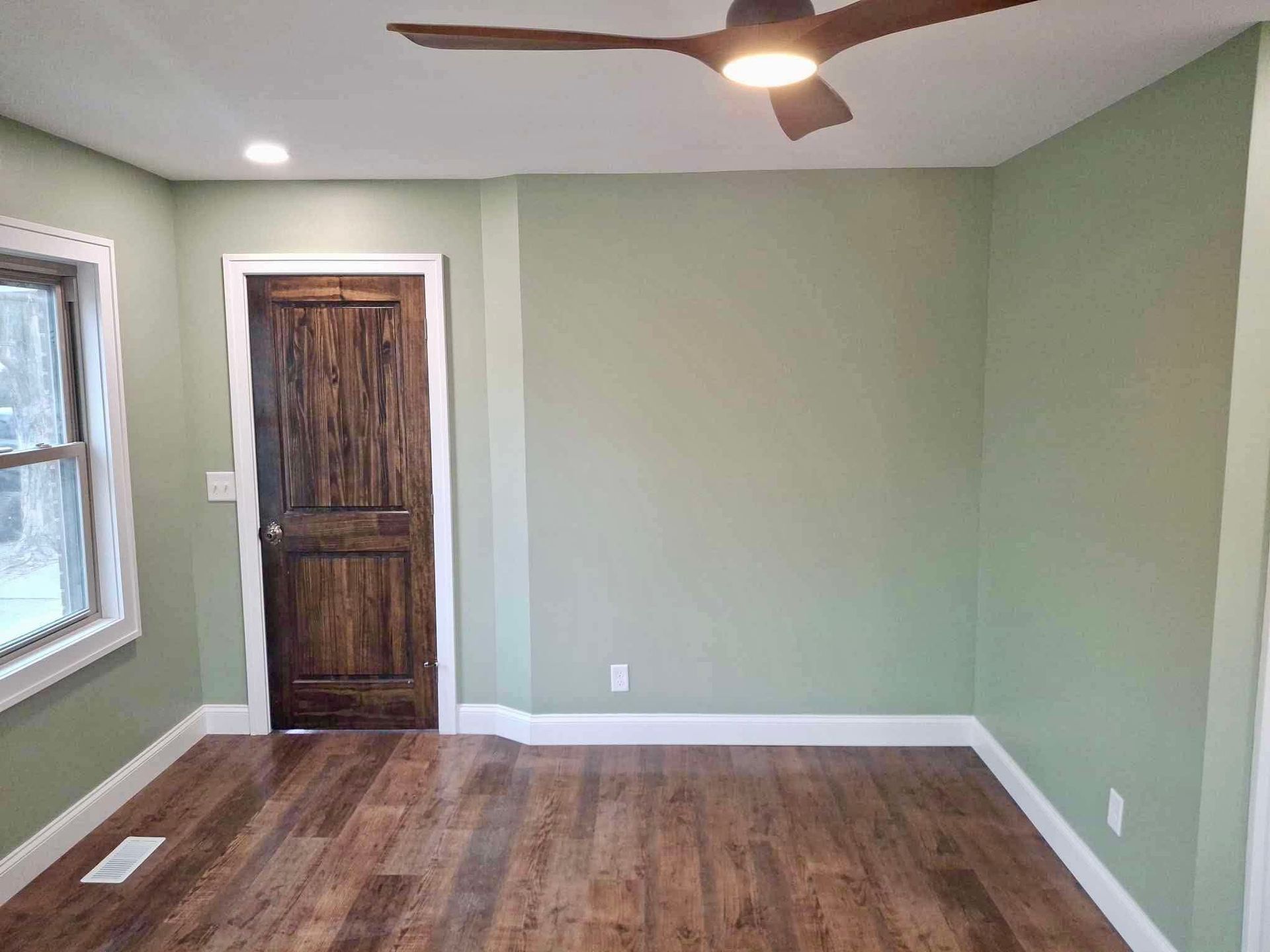 Empty room with light green walls, wood floor, ceiling fan, window, and dark wood door.
