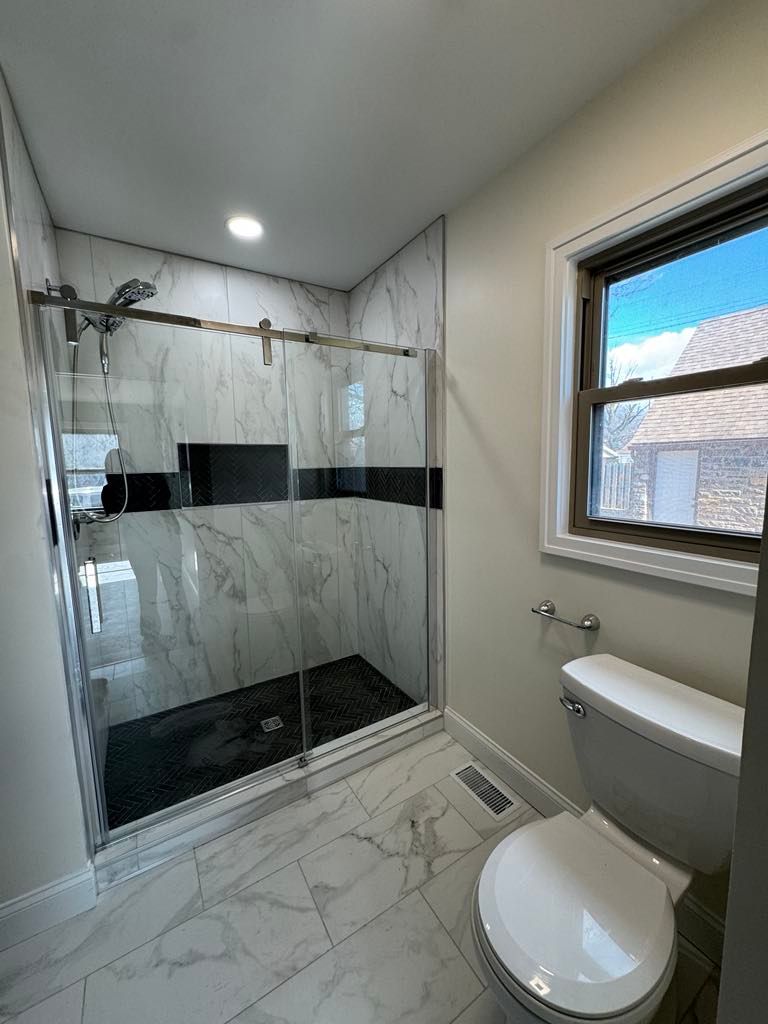 Modern bathroom with marble shower, glass doors, and a toilet beside a window.