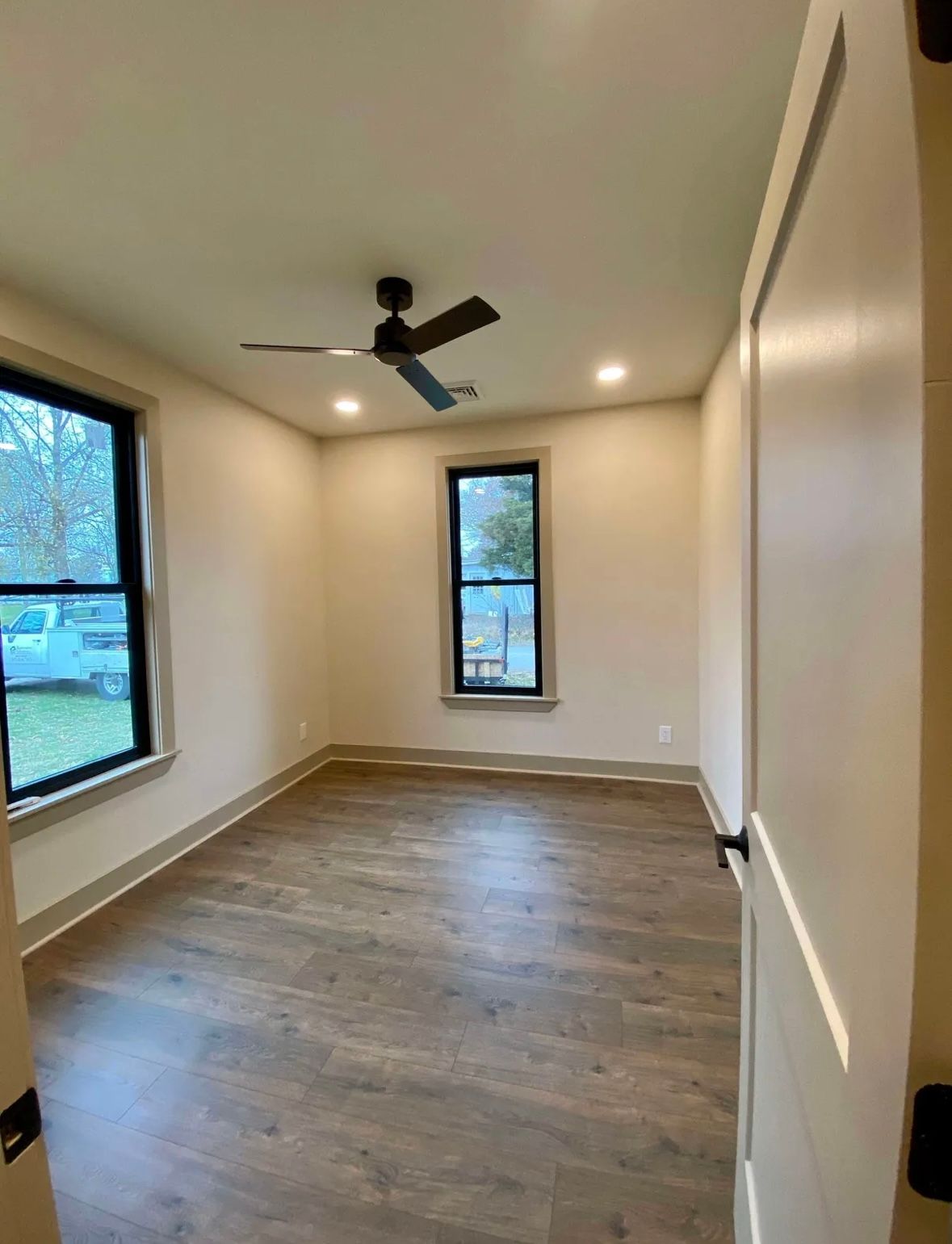 Empty narrow room with wood floors, cream walls, a ceiling fan, and two windows letting in daylight