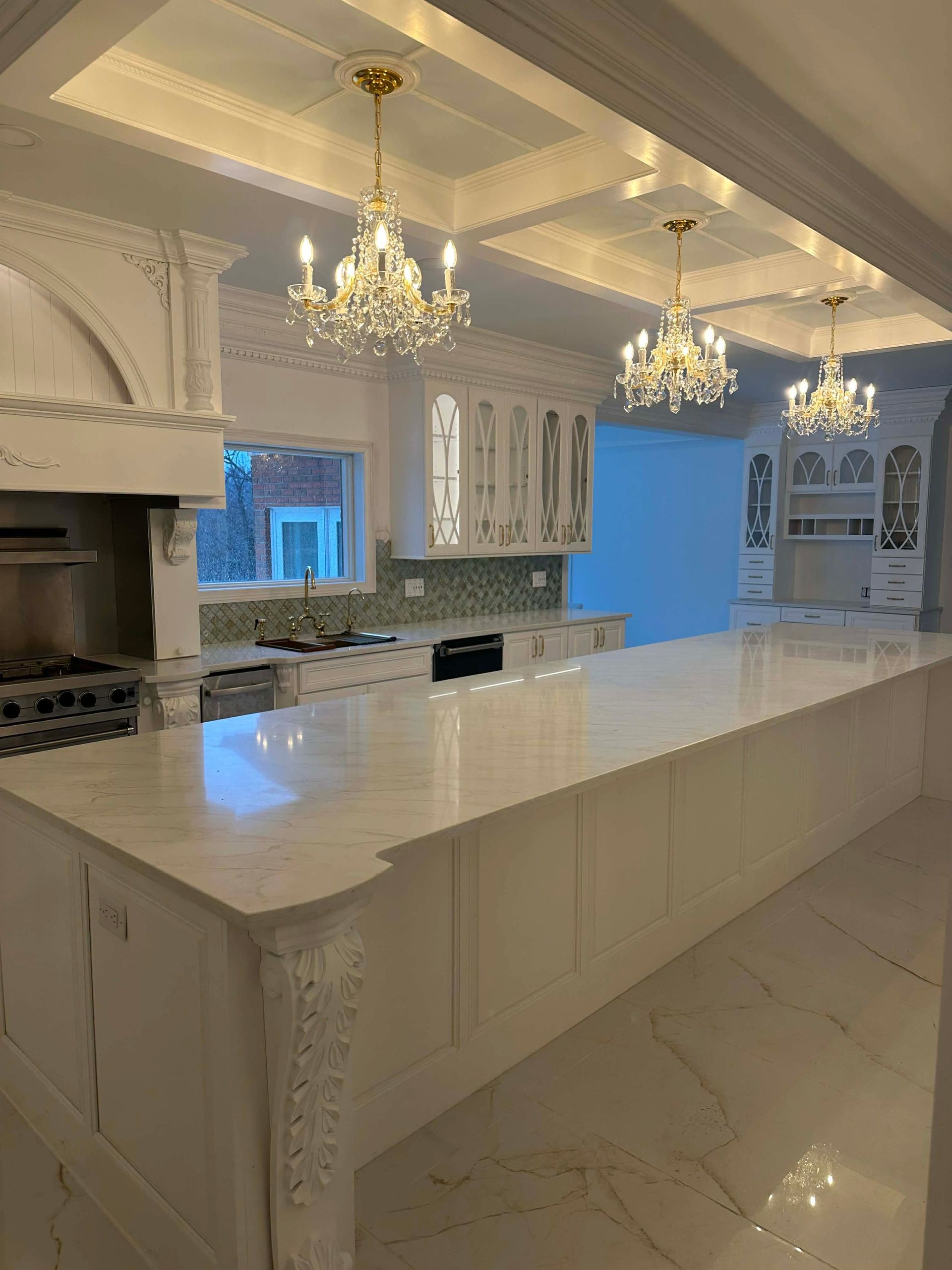 Elegant white kitchen with marble island, chandeliers, ornate cabinets, and a stove alcove.