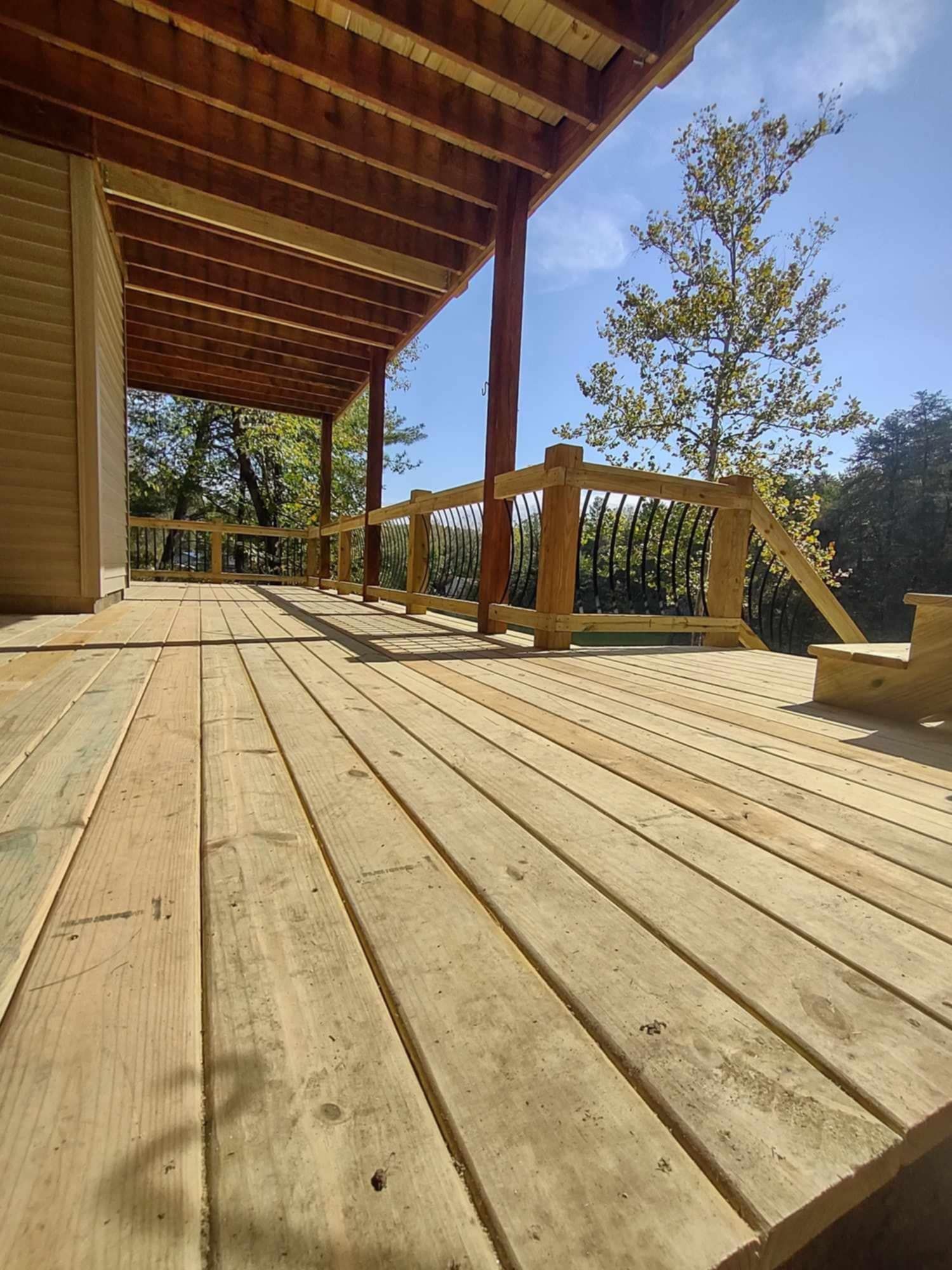 Wooden deck with railing and roof overhang, overlooking trees in bright sunlight.