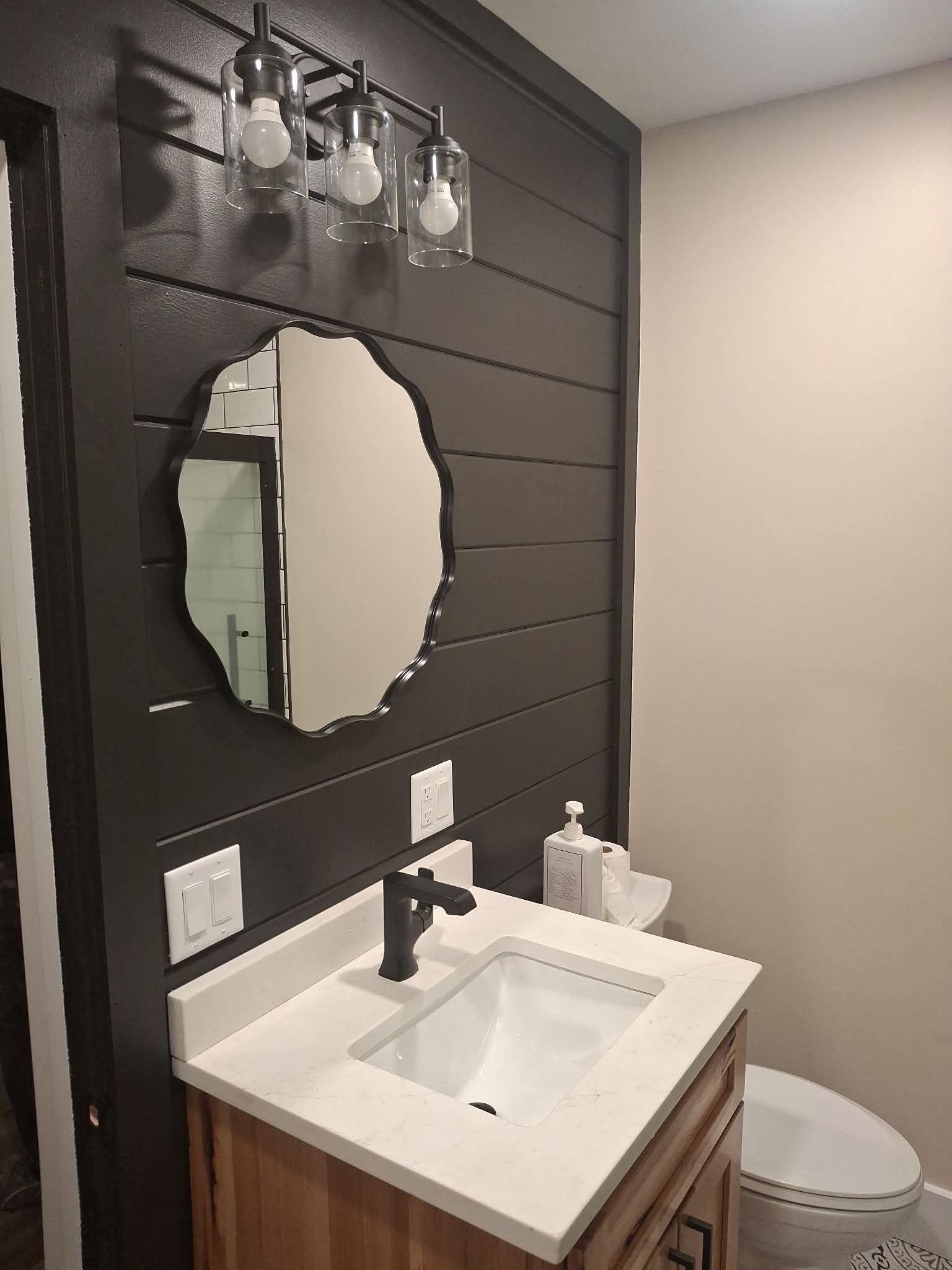 Small bathroom vanity with white sink, black faucet, oval mirror, and toilet against dark wood paneling.