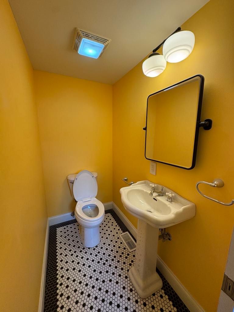 Small yellow bathroom with toilet, pedestal sink, mirror, and overhead light.