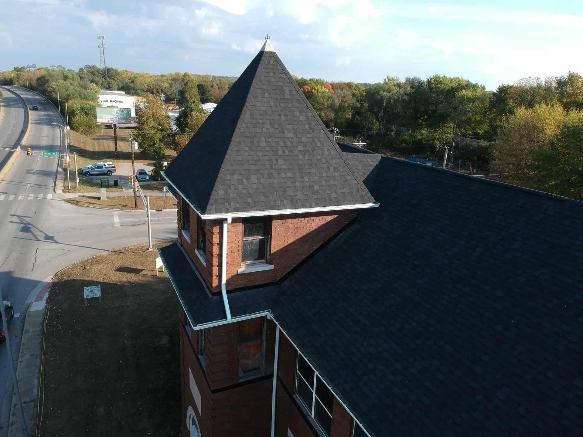 Brick building with dark shingled roof and tower, viewed from above near a road and trees