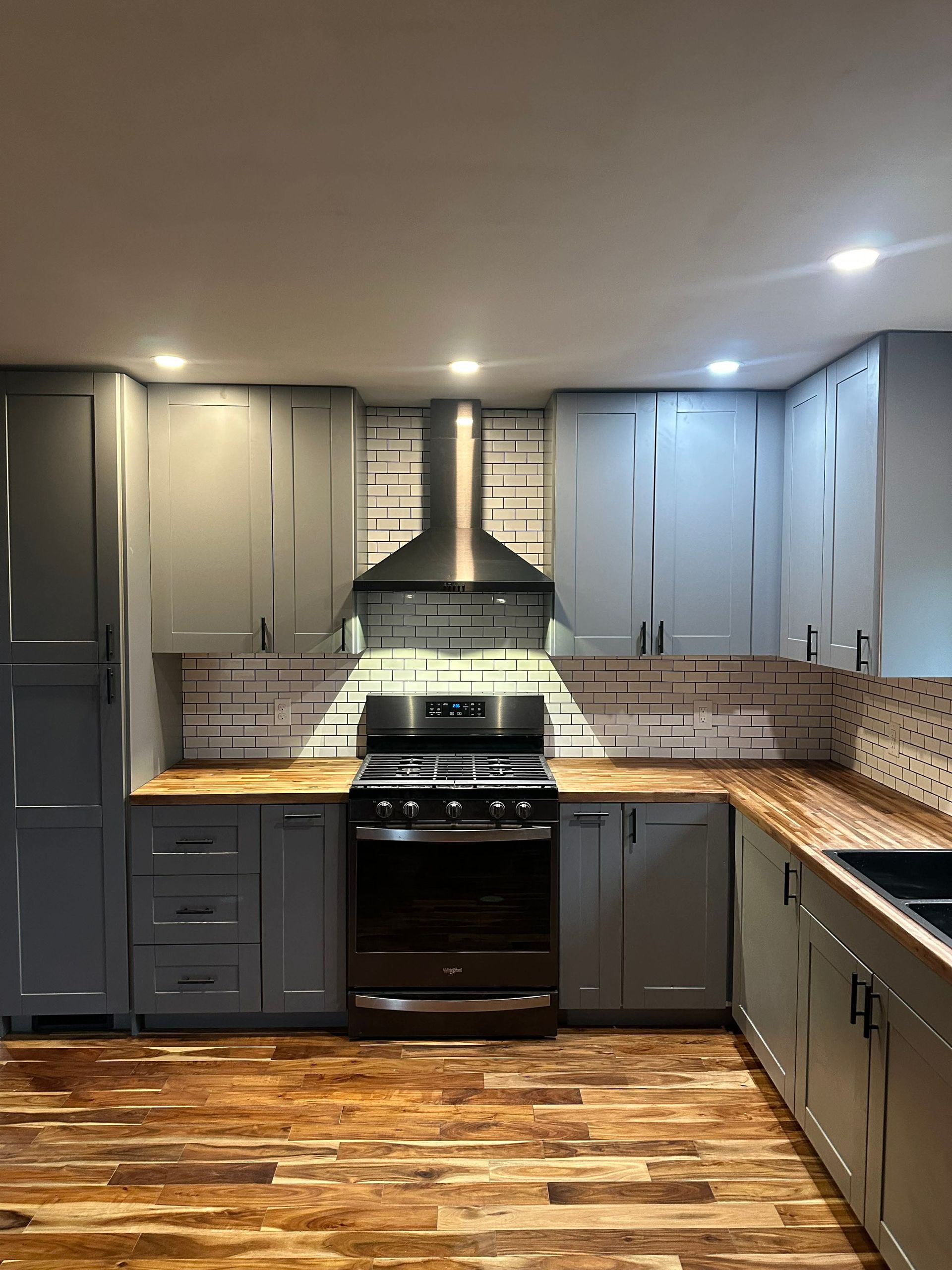 Modern kitchen with gray cabinets, black stove, stainless hood, and wood floor.