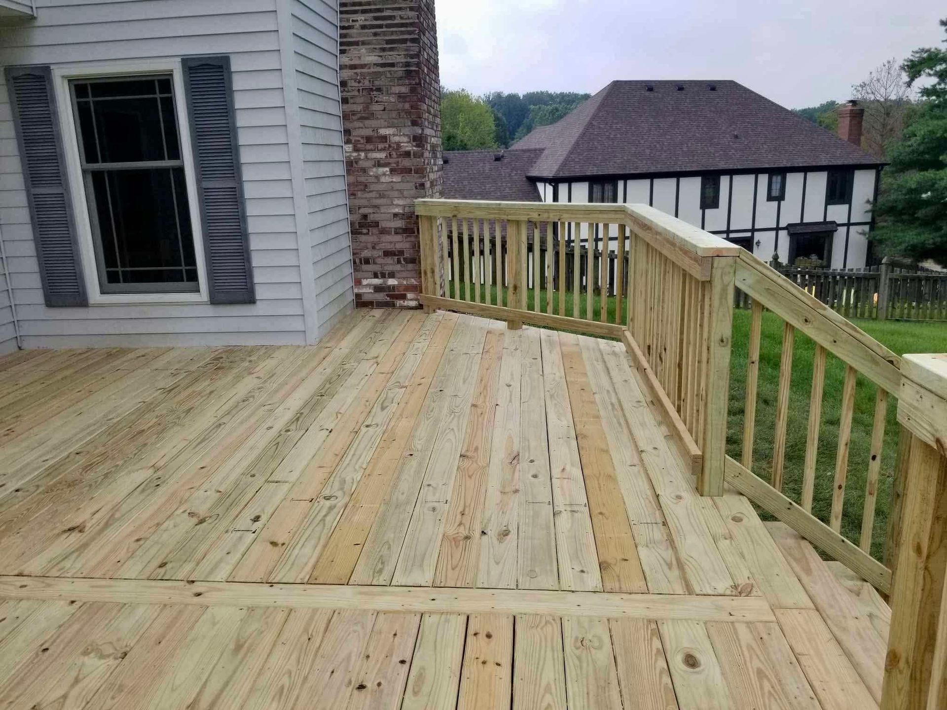 Wooden deck beside a house, with railing and stairs overlooking a yard and neighboring homes.