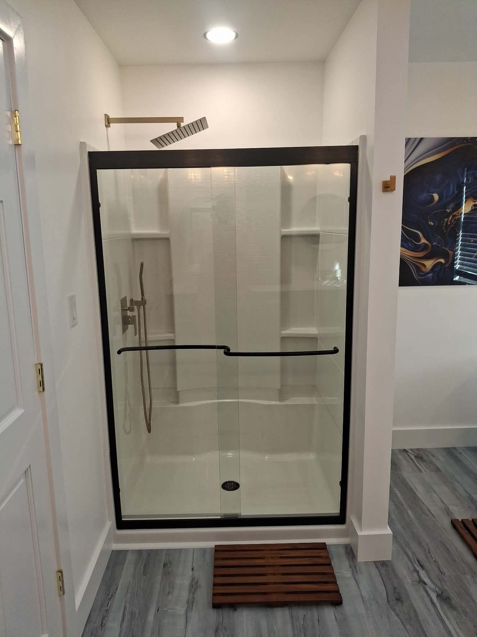 Glass shower enclosure in a small white bathroom with a bath mat in front and wall art on the right.