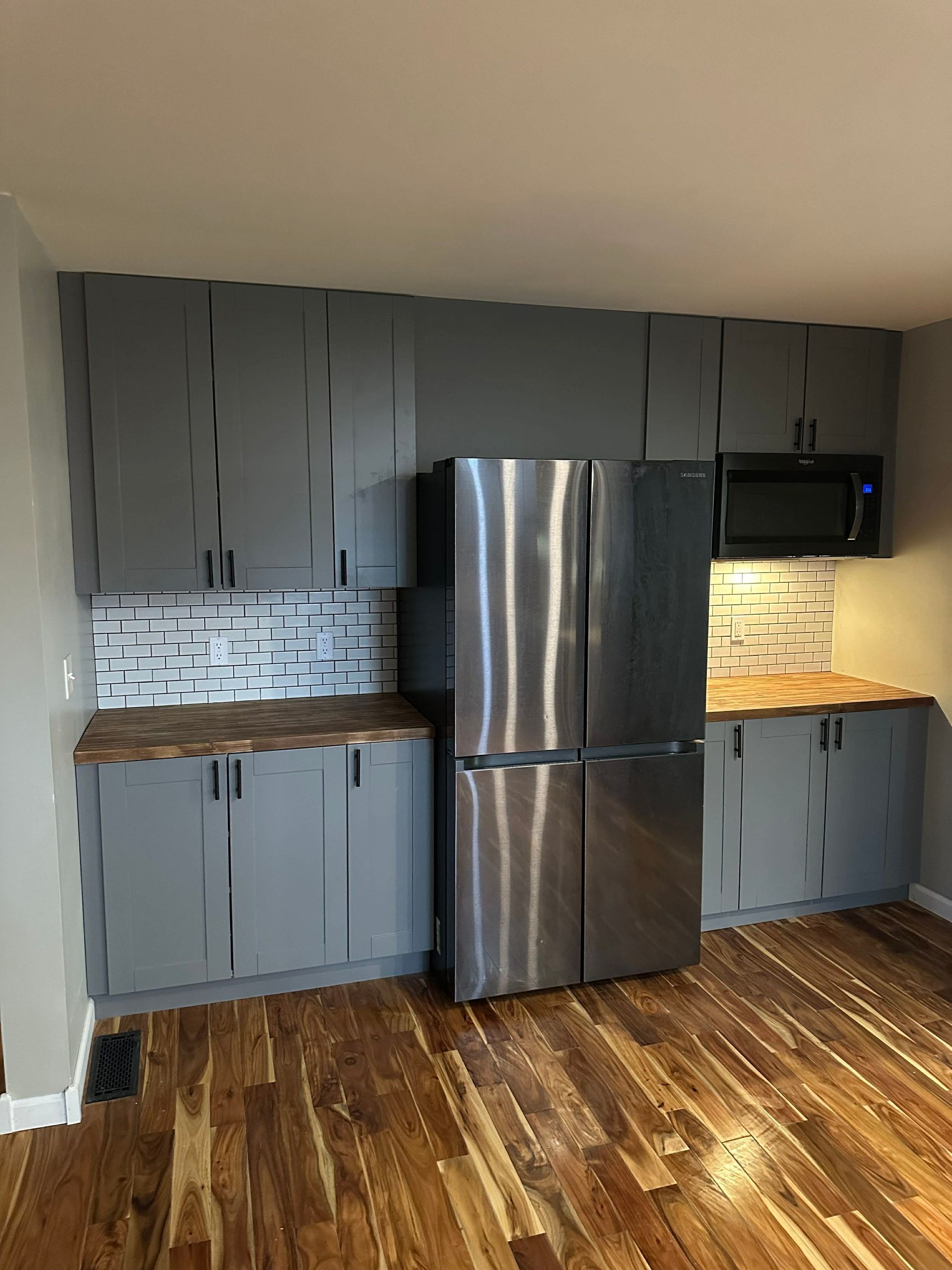 Small kitchen with gray cabinets, stainless steel fridge, black microwave, and wood floors.