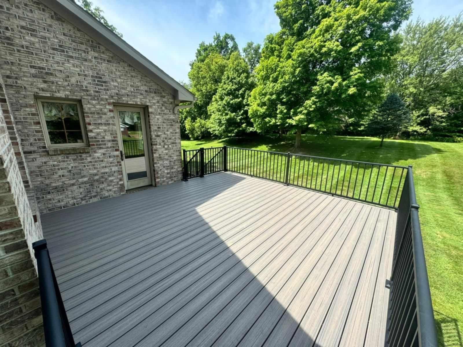 Spacious gray composite deck beside a stone house, bordered by black railing and overlooking a green lawn and trees