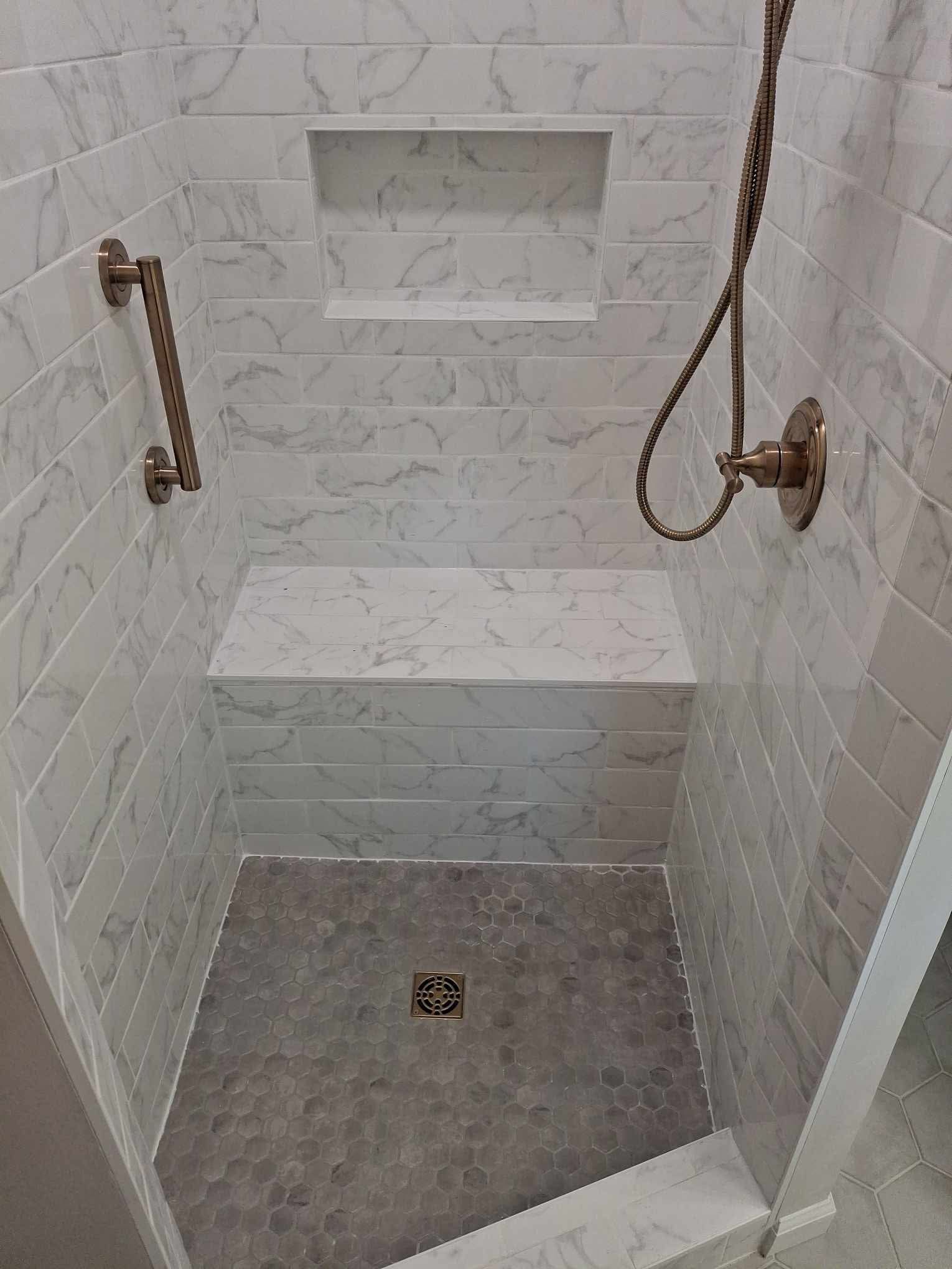 Small white tiled shower stall with built-in bench, recessed shelf, grab bars, and floor drain