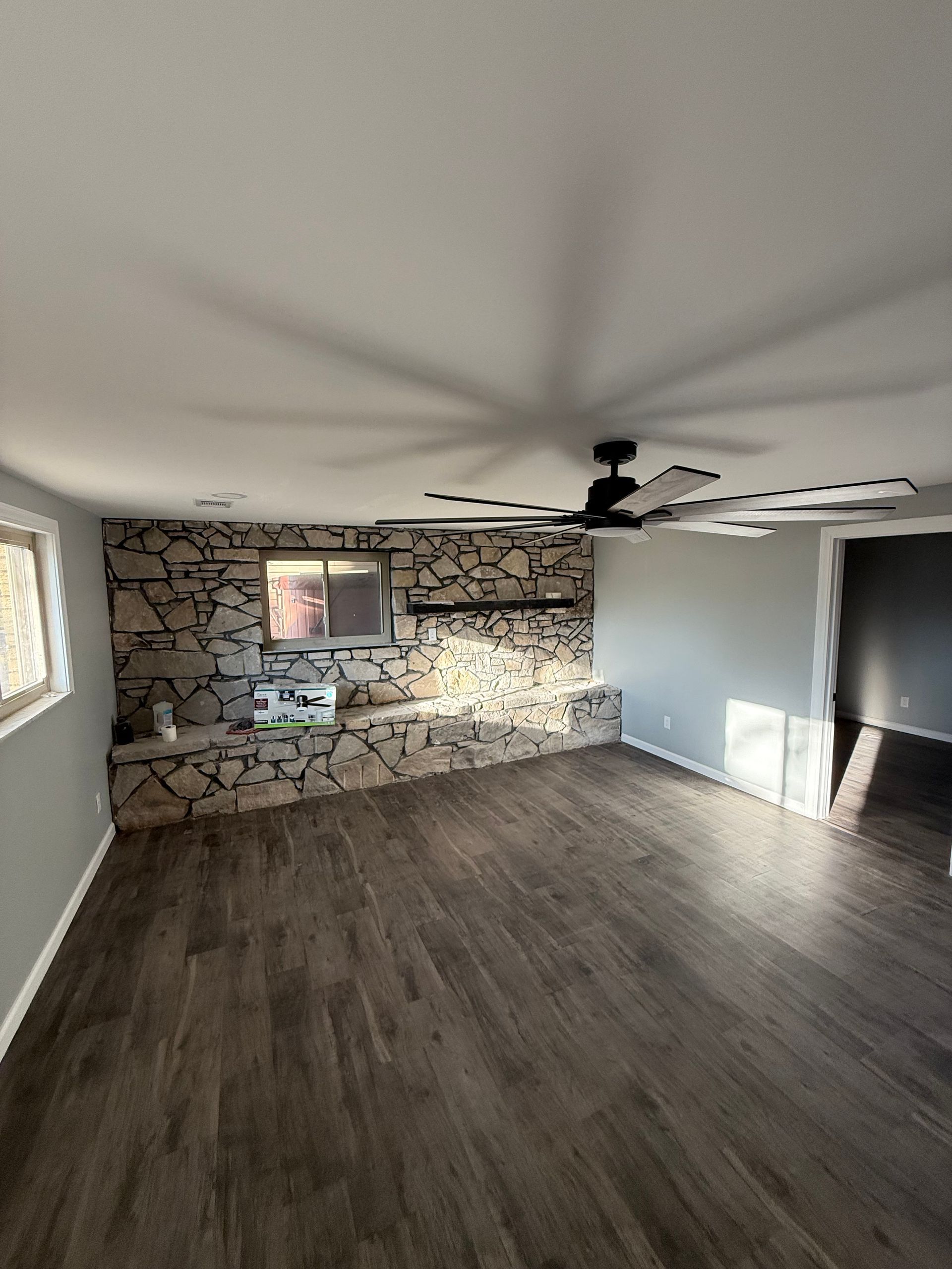 Empty living room with stone accent wall, gray wood floor, ceiling fan, and bright windows