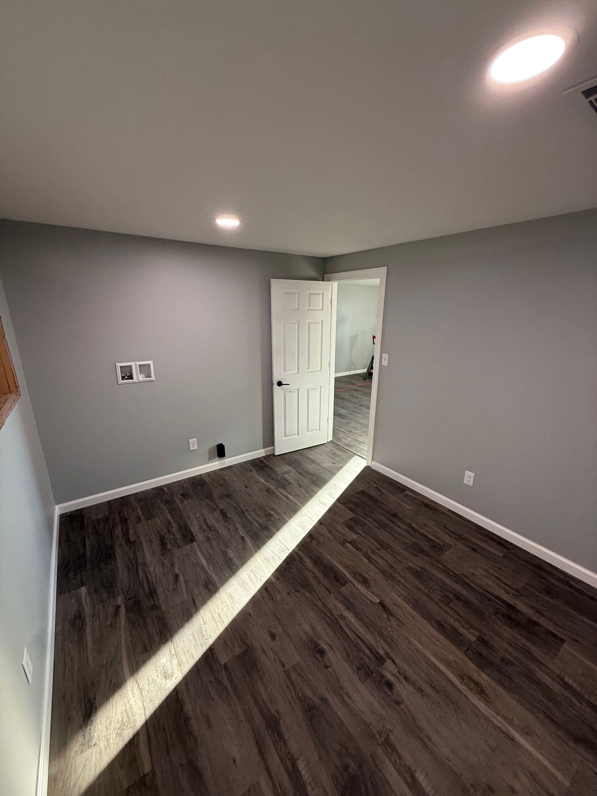 Empty gray bedroom with dark wood floors, white trim, and an open door to a lit hallway