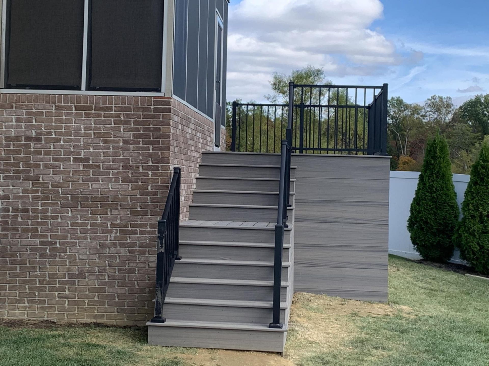 Exterior stairs with a black railing leading to a brick building and deck, beside a grassy yard.