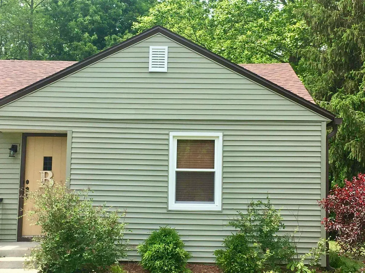 Small green house exterior with front door, window, and shrubs in front