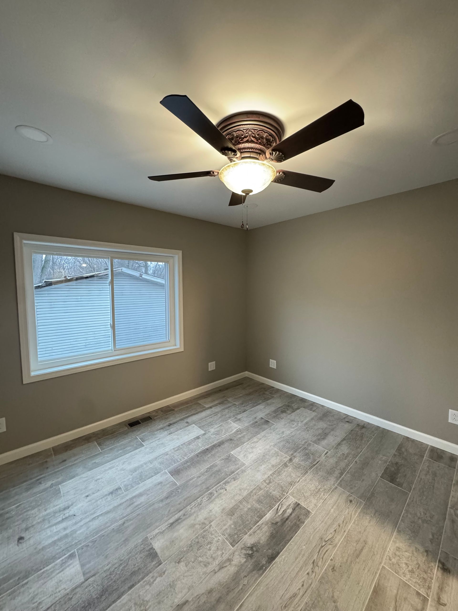 Empty bedroom with gray walls, wood-look flooring, a window, and a ceiling fan with lights.
