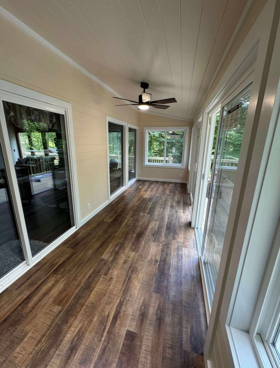 Long sunroom with wood-look floor, white walls, ceiling fan, and large windows overlooking greenery