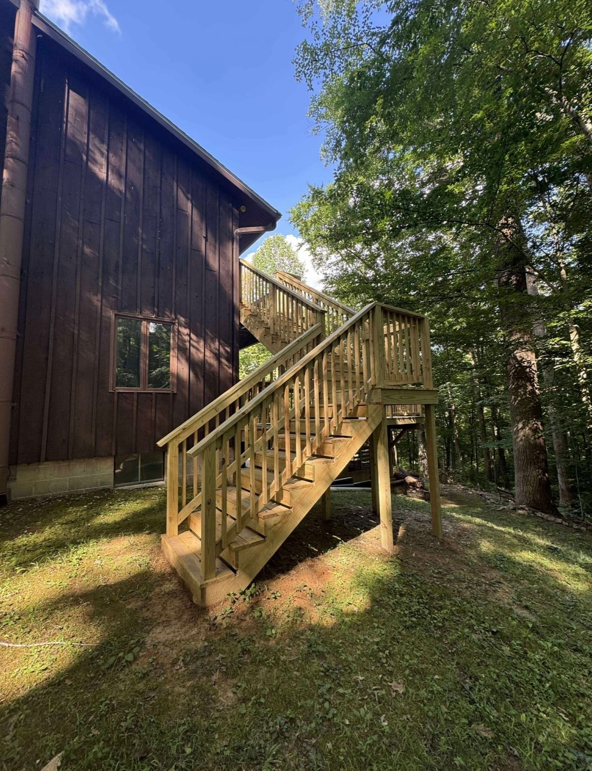 Wooden exterior staircase beside a dark brown house, with trees and grassy yard in sunlight.