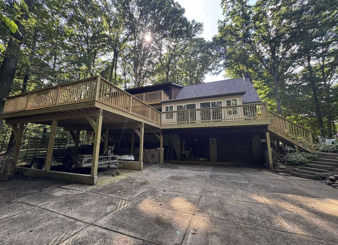 Wooded hillside house with wraparound deck, gravel driveway, and parked car under trees