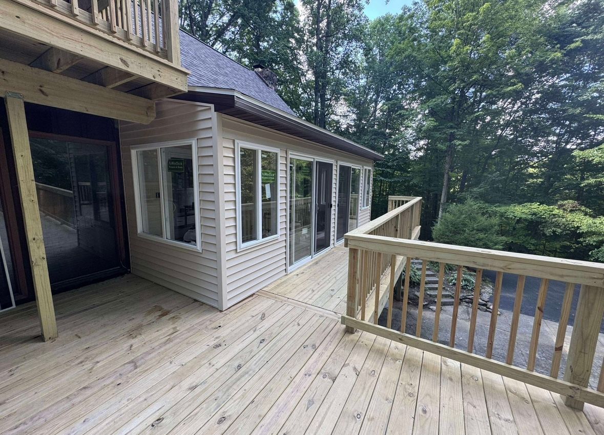 Wooden deck beside a house with white siding, railing, and trees beyond.