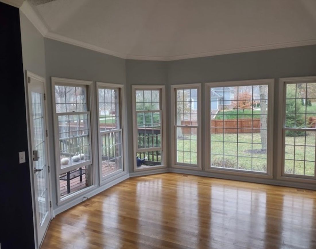 Bright empty sunroom with bay windows, gray walls, and hardwood floors overlooking a backyard.