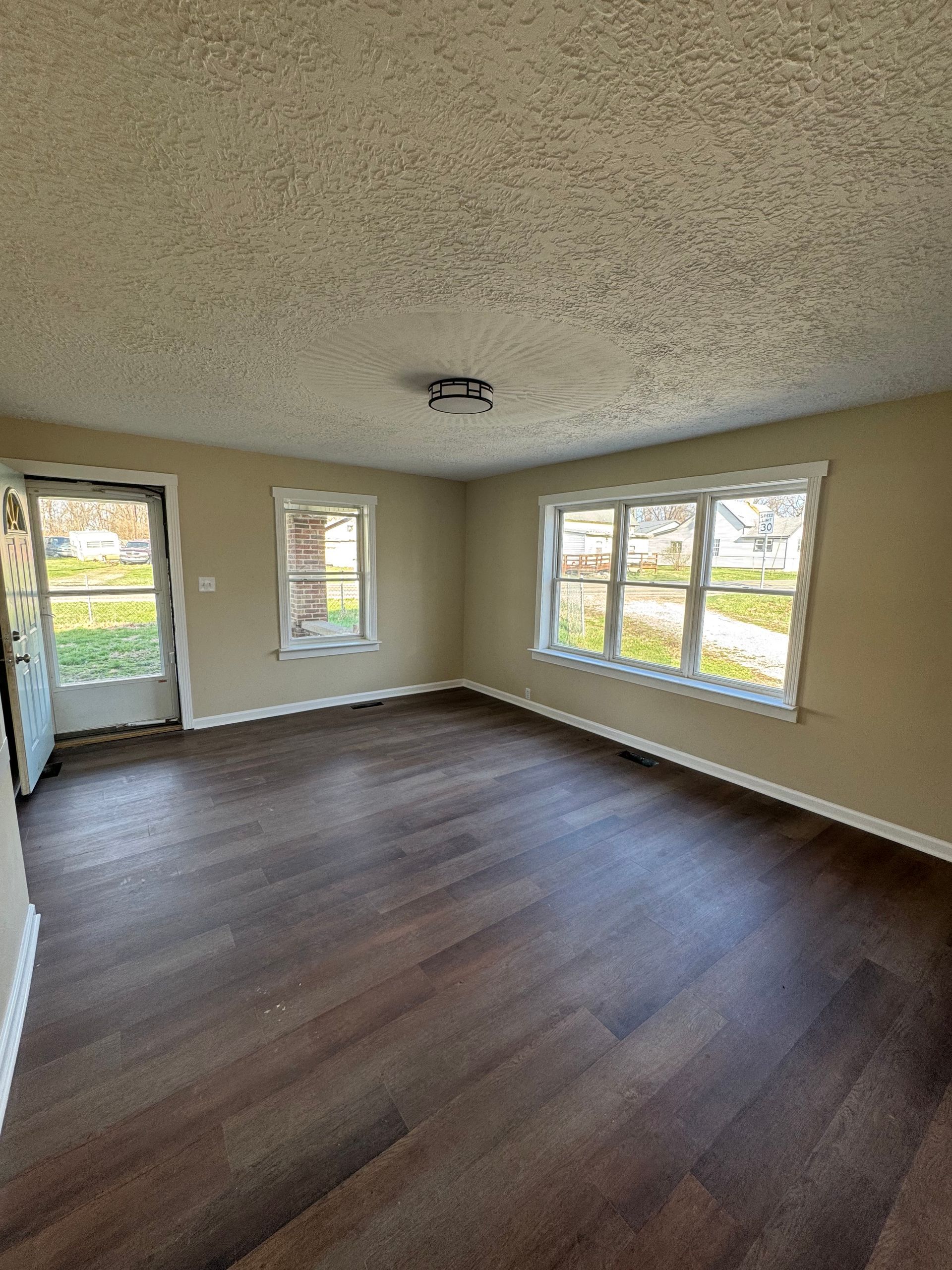 Empty living room with dark wood floors, beige walls, and large windows overlooking a yard