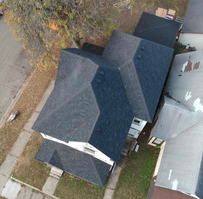 Aerial view of a dark-shingled house with a small yard and neighboring roofs.