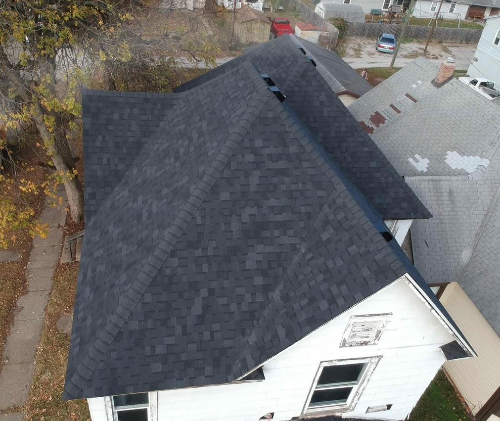 Aerial view of a house with a dark shingled roof and white siding in a residential neighborhood