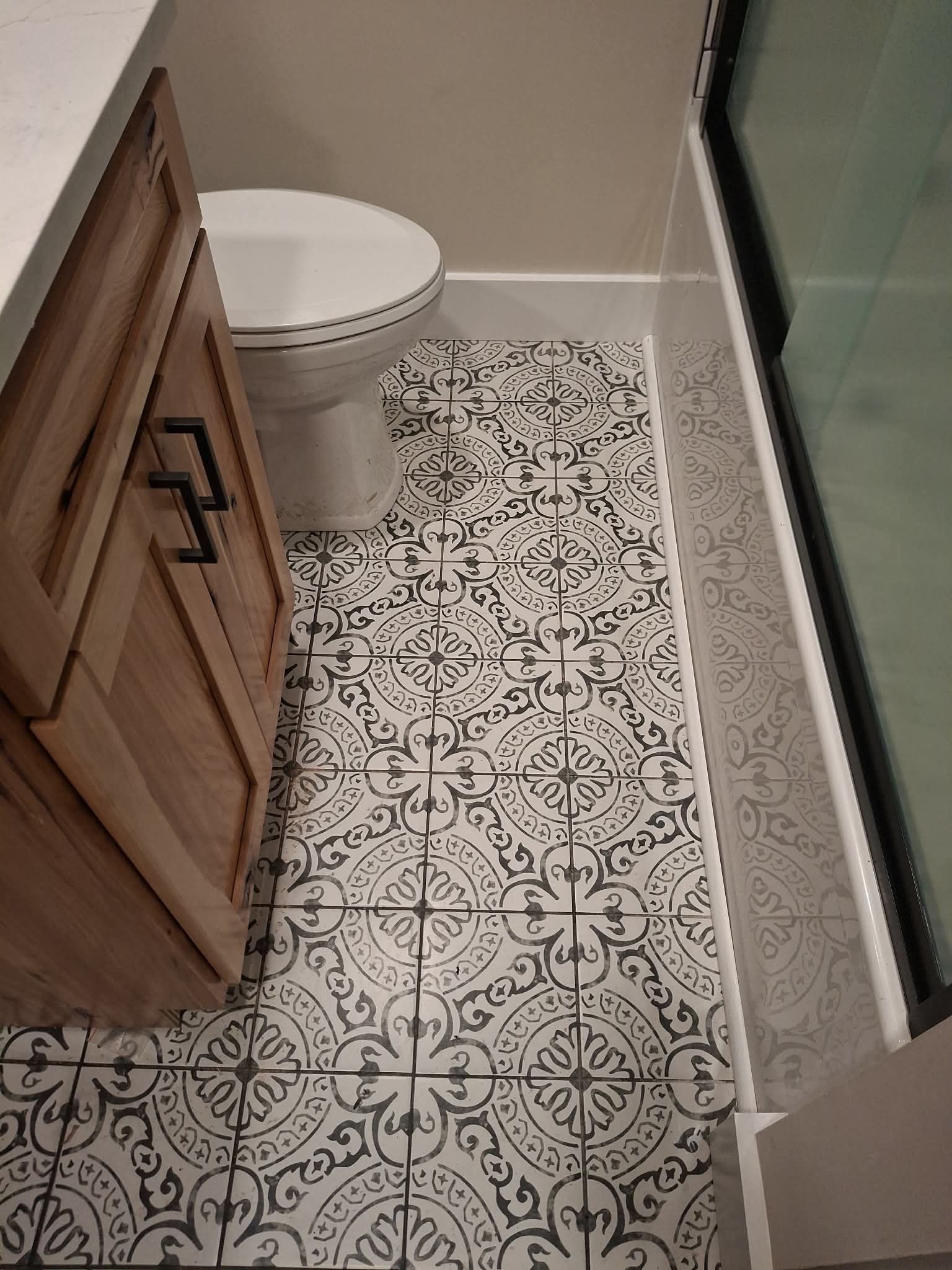 Narrow bathroom with patterned tile floor, toilet, wooden vanity, and glass shower door.