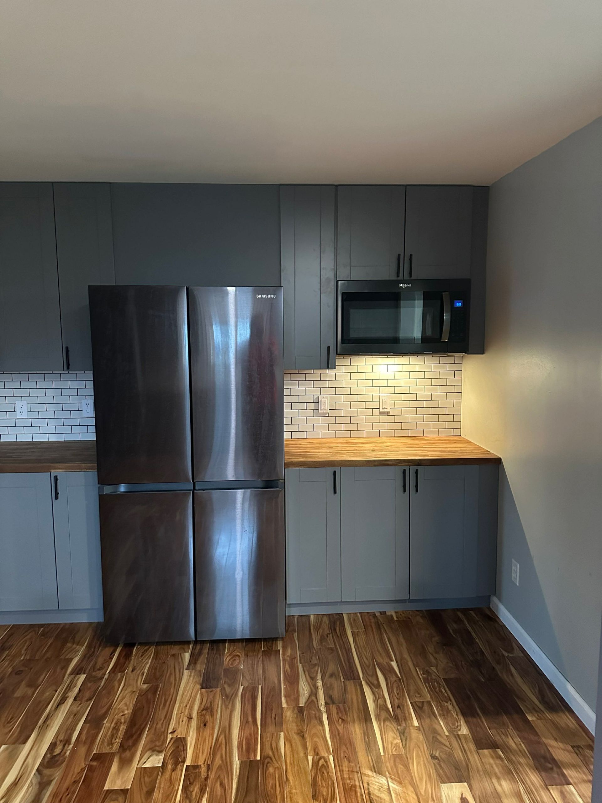 Empty kitchen with dark cabinets, stainless steel fridge, microwave, and warm under-cabinet lighting over wood floors