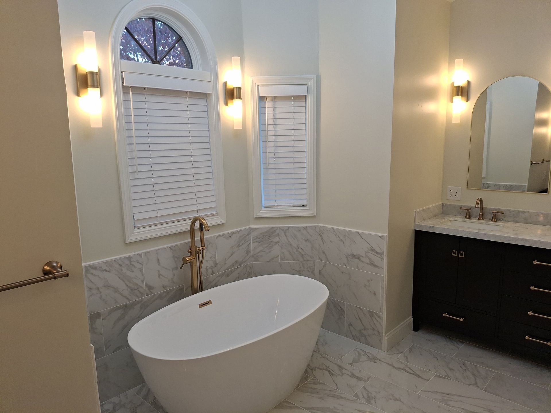 Modern bathroom with freestanding tub, marble tile, double vanity, and arched window blinds