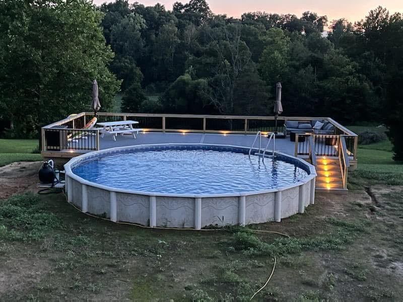 Above-ground backyard pool with wooden deck, lit steps, and trees at dusk