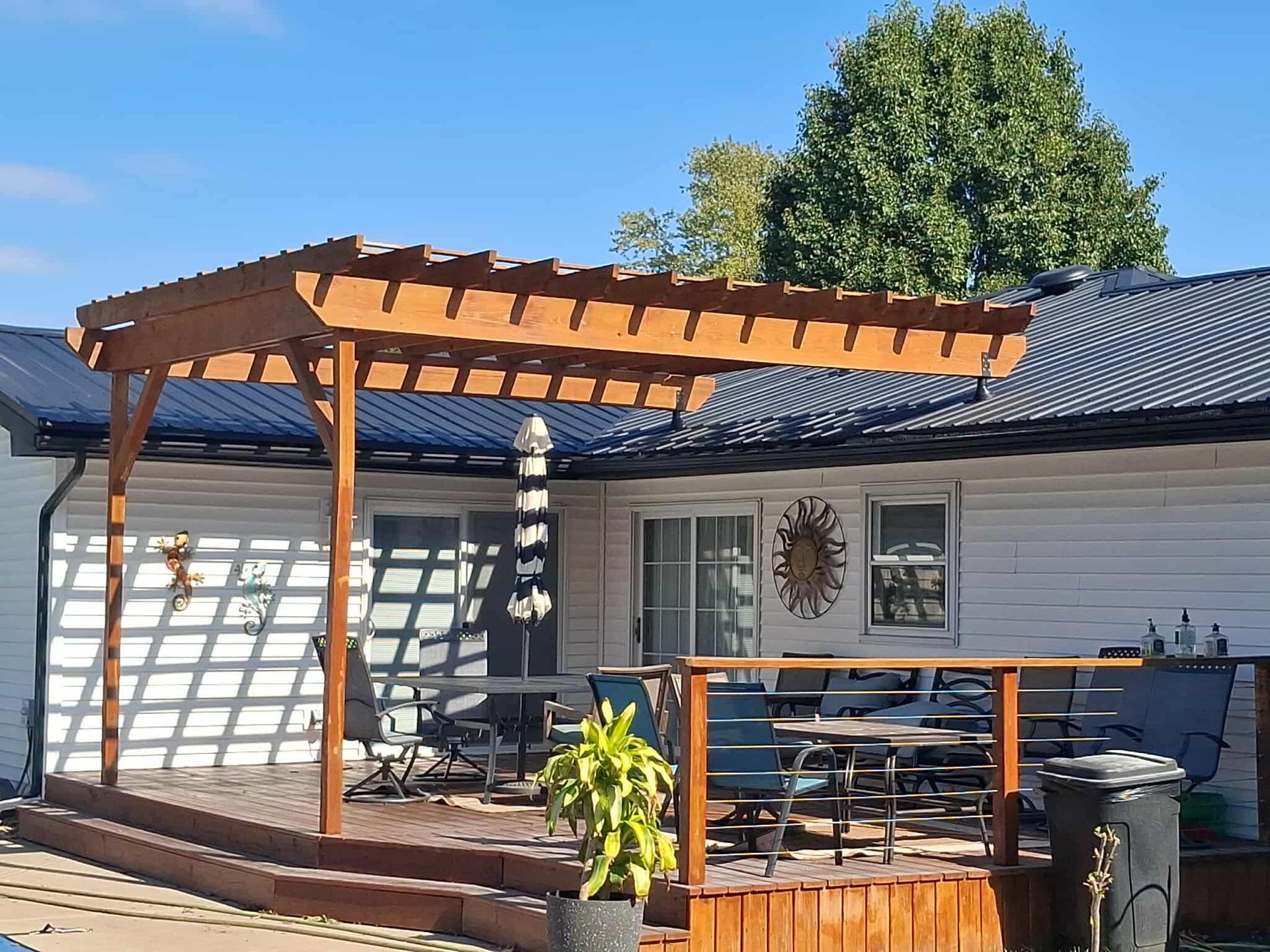 Backyard deck with wooden pergola, railing, table, chairs, and a house exterior under a clear blue sky