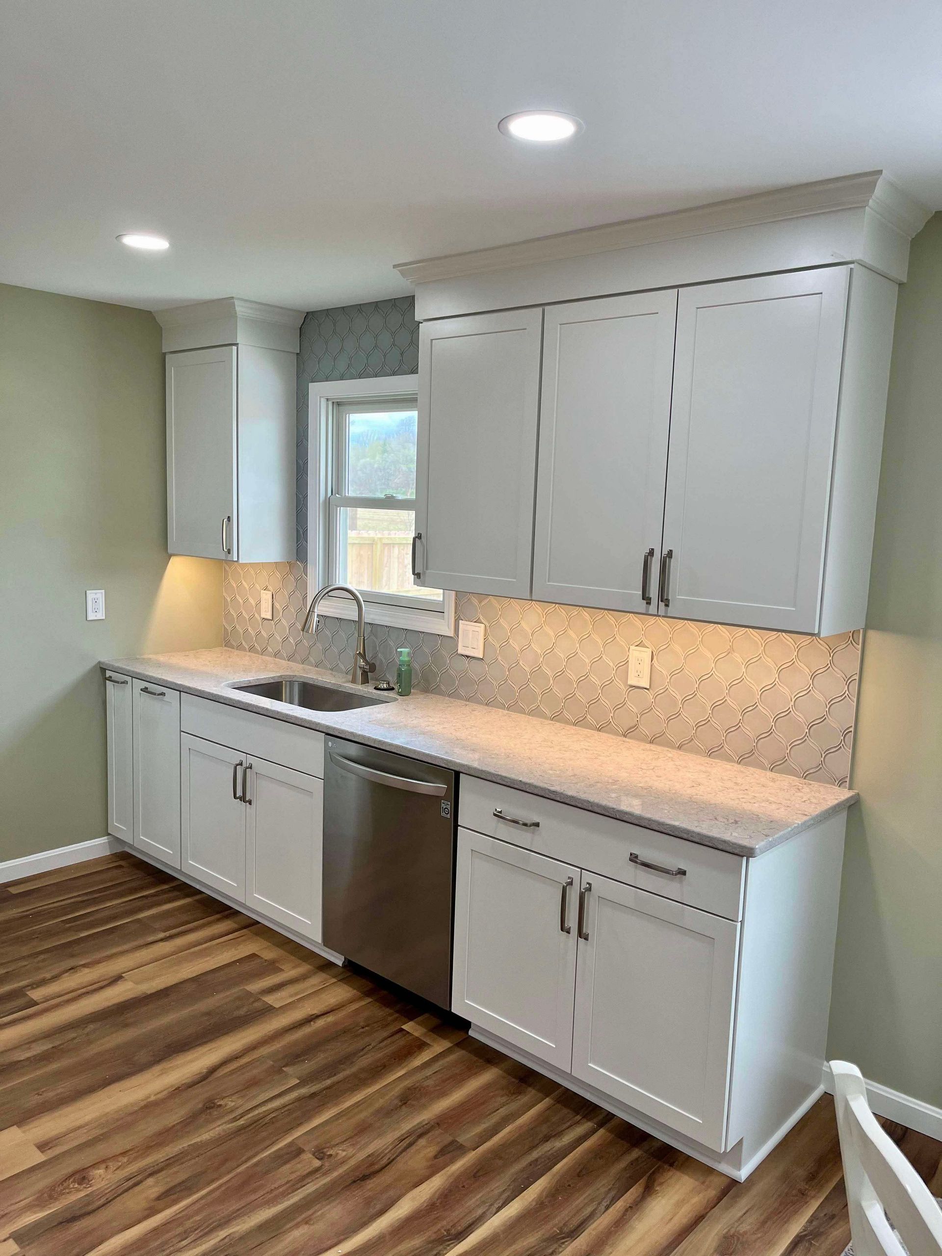 Modern kitchen with white cabinets, stainless dishwasher, sink under window, and wood-look flooring