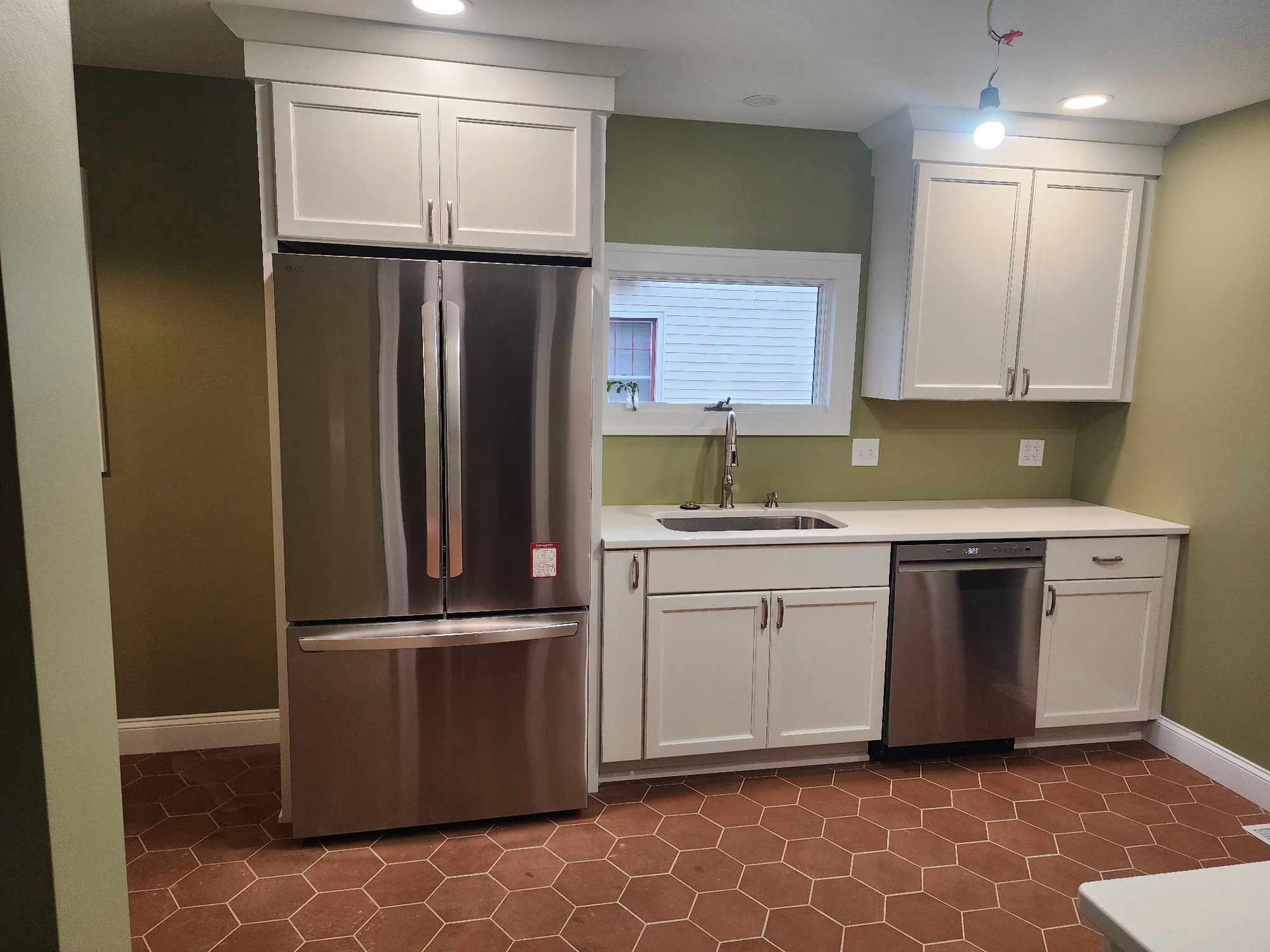 Modern kitchen with stainless steel fridge, white cabinets, window, sink, and dishwasher on red tile floor