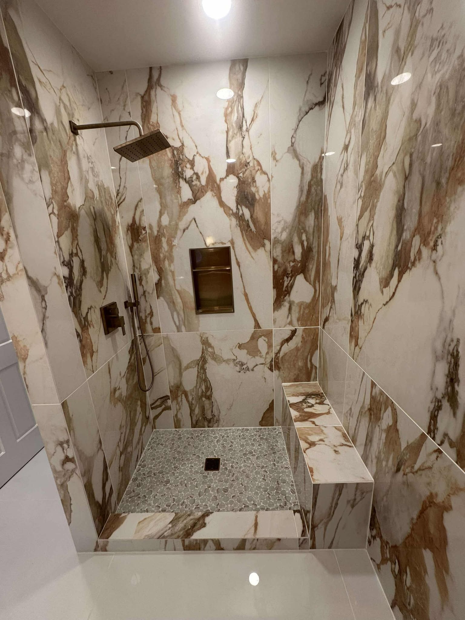 Marble-tiled walk-in shower with a pebble floor, built-in bench, and brass fixtures
