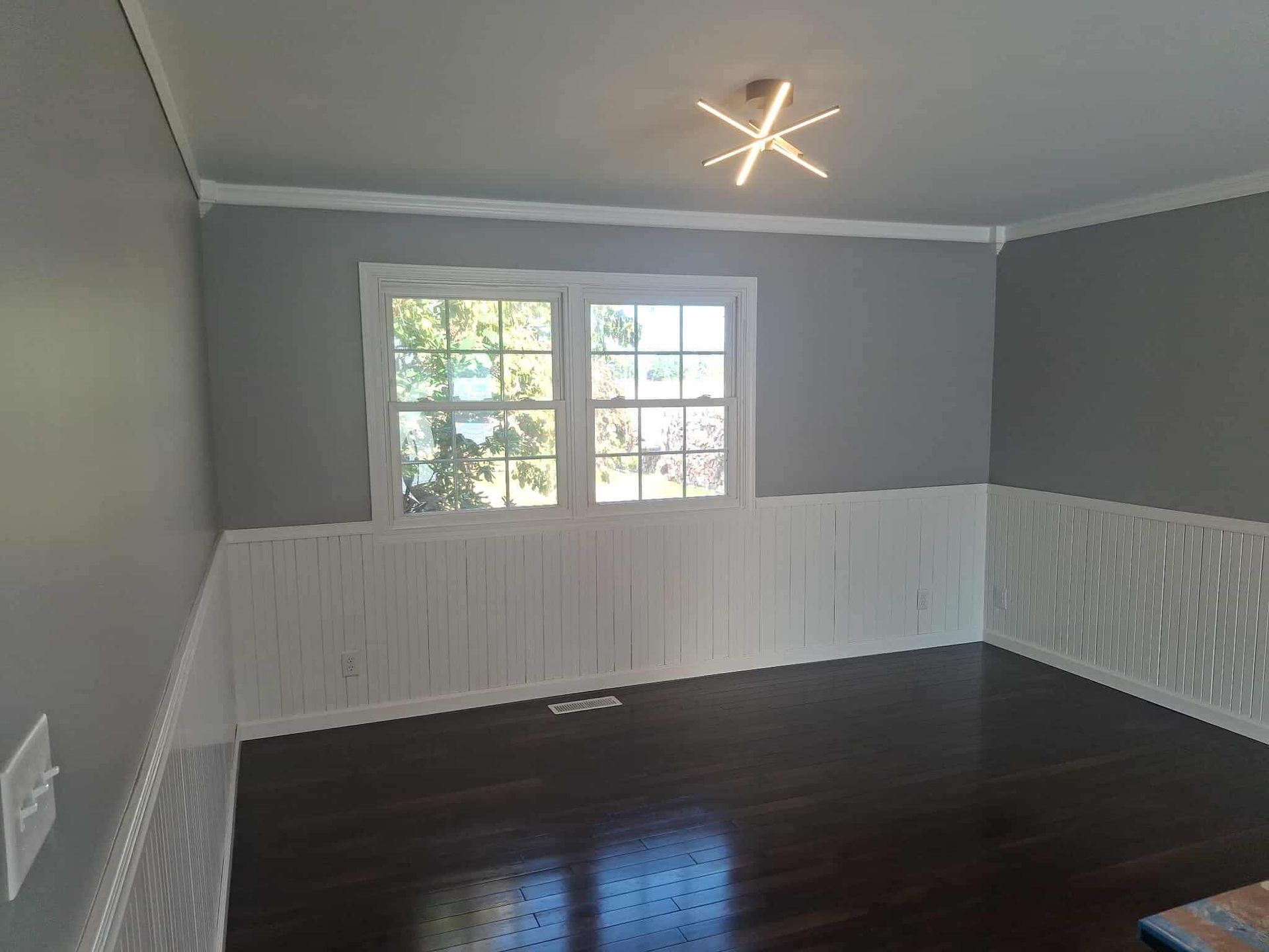 Empty room with dark hardwood floors, gray walls, white wainscoting, and a ceiling light near a window.