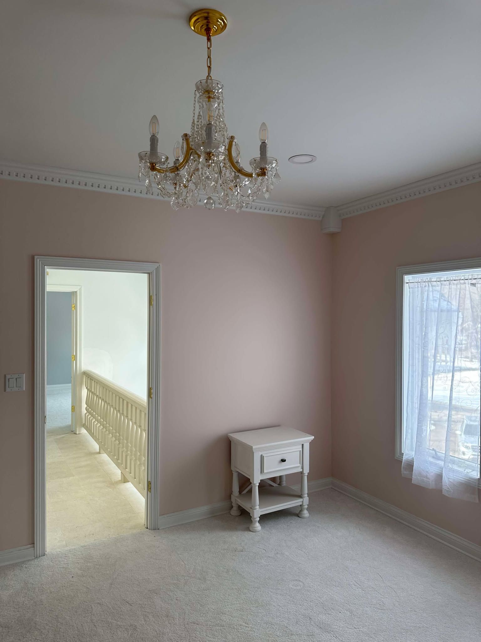 Empty pink bedroom with chandelier, carpet, doorway to hall, and a small white table by a window.