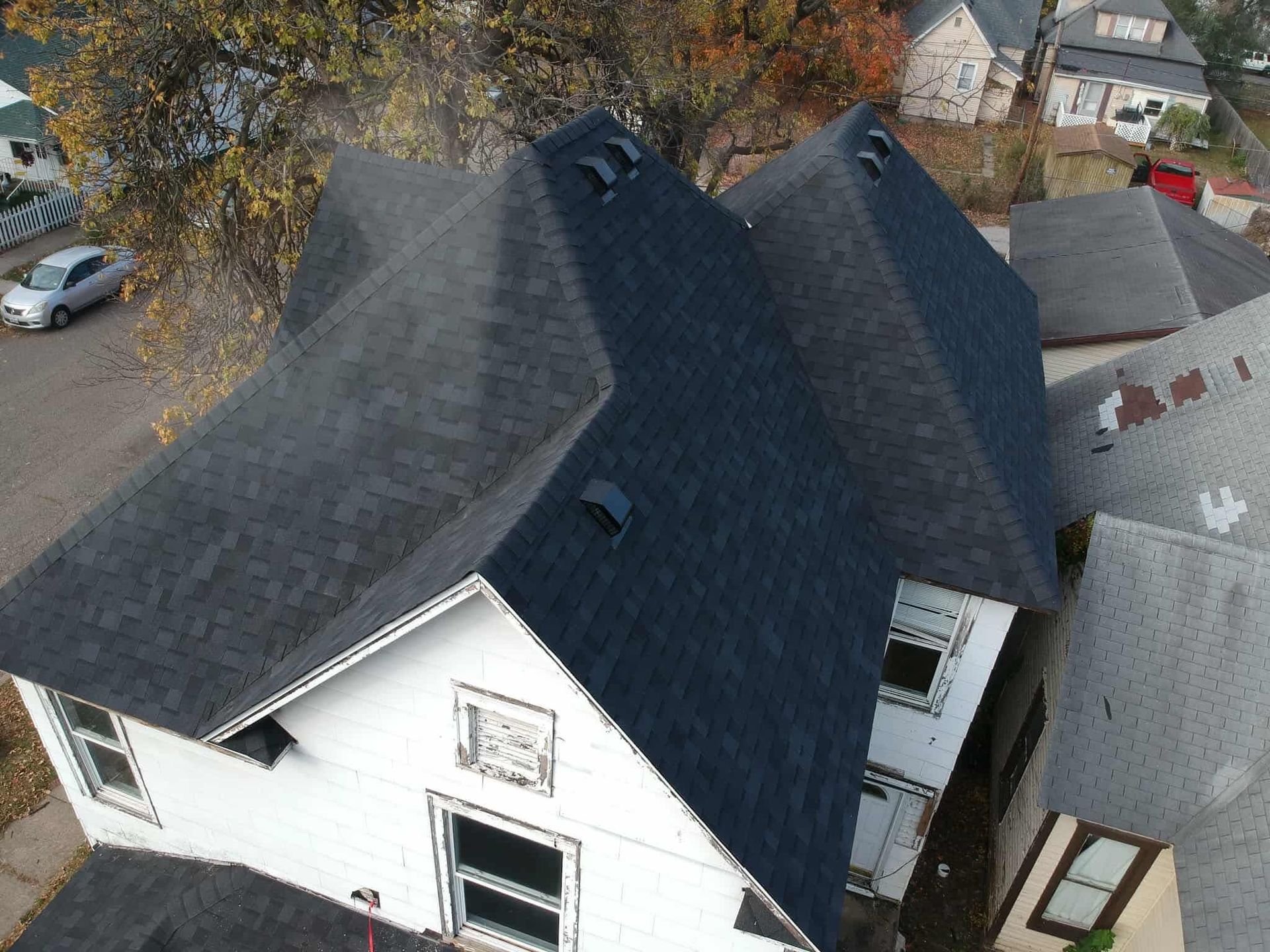 Aerial view of a white house with a dark shingled roof and nearby trees in autumn.