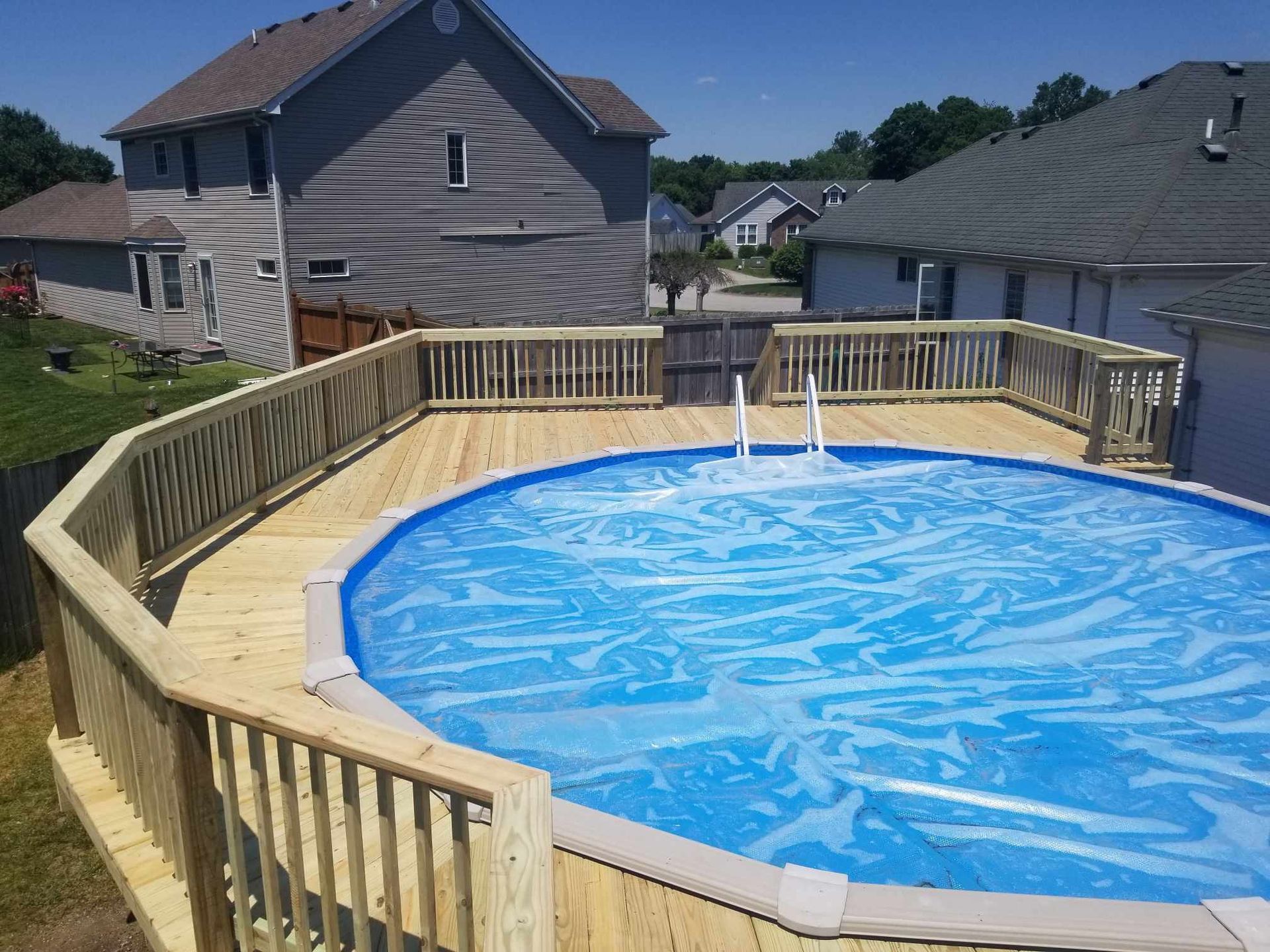 Above-ground pool with blue water on a wooden deck in a suburban backyard.