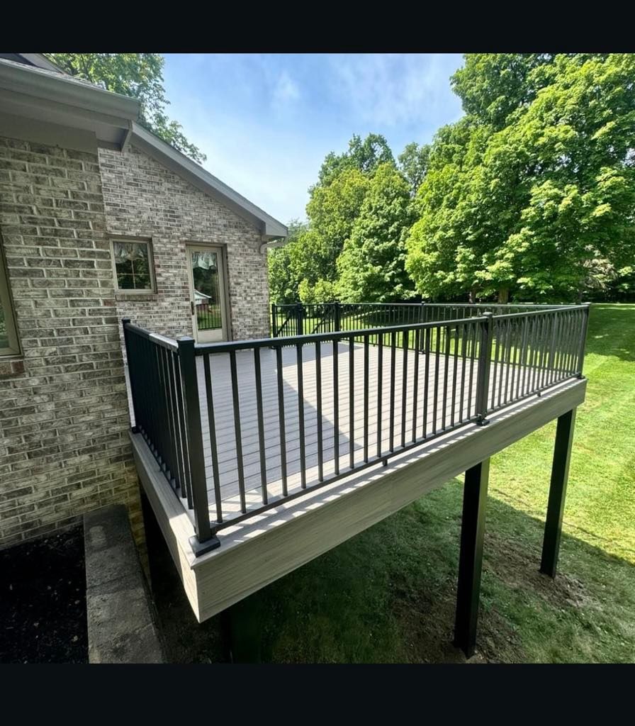 Raised wooden deck with black railings beside a stone house, overlooking a grassy yard.