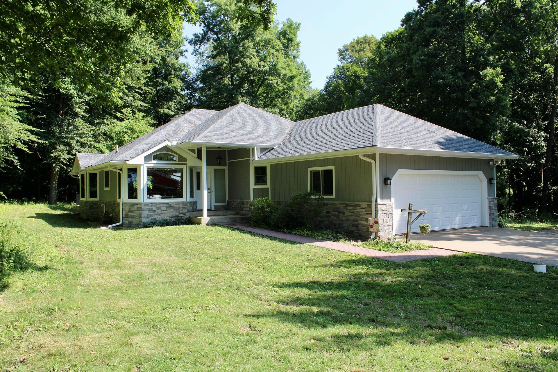 Single-story green house with front porch, white garage, and large grassy yard surrounded by trees