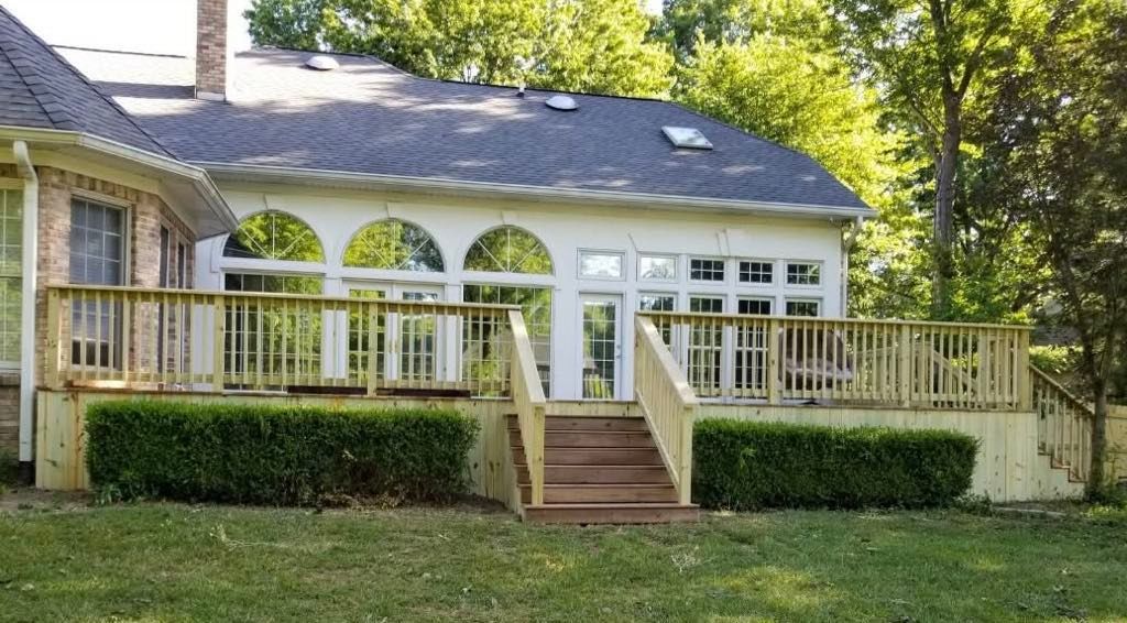 Front view of a white house with a wooden porch, steps, and green hedges under trees