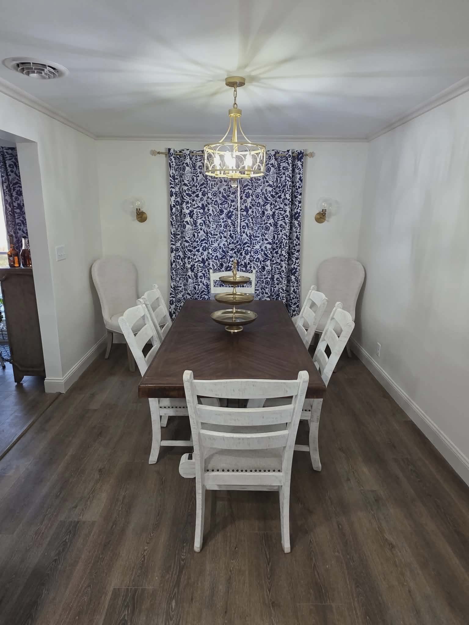 Dining room with dark wood table, white chairs, chandelier, and patterned blue curtains.