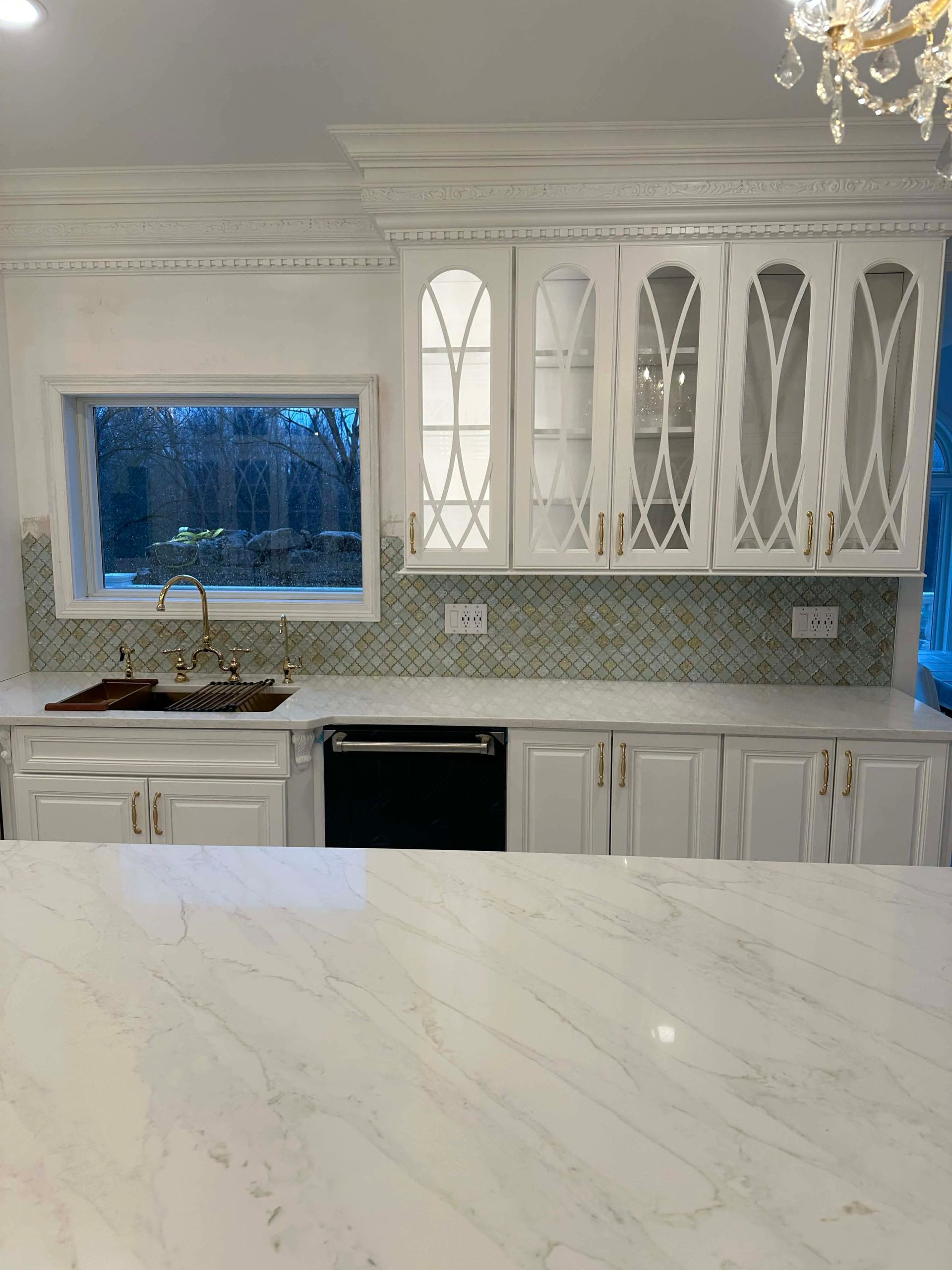 Bright white kitchen with marble island, glass cabinets, chandelier, and window above the sink.