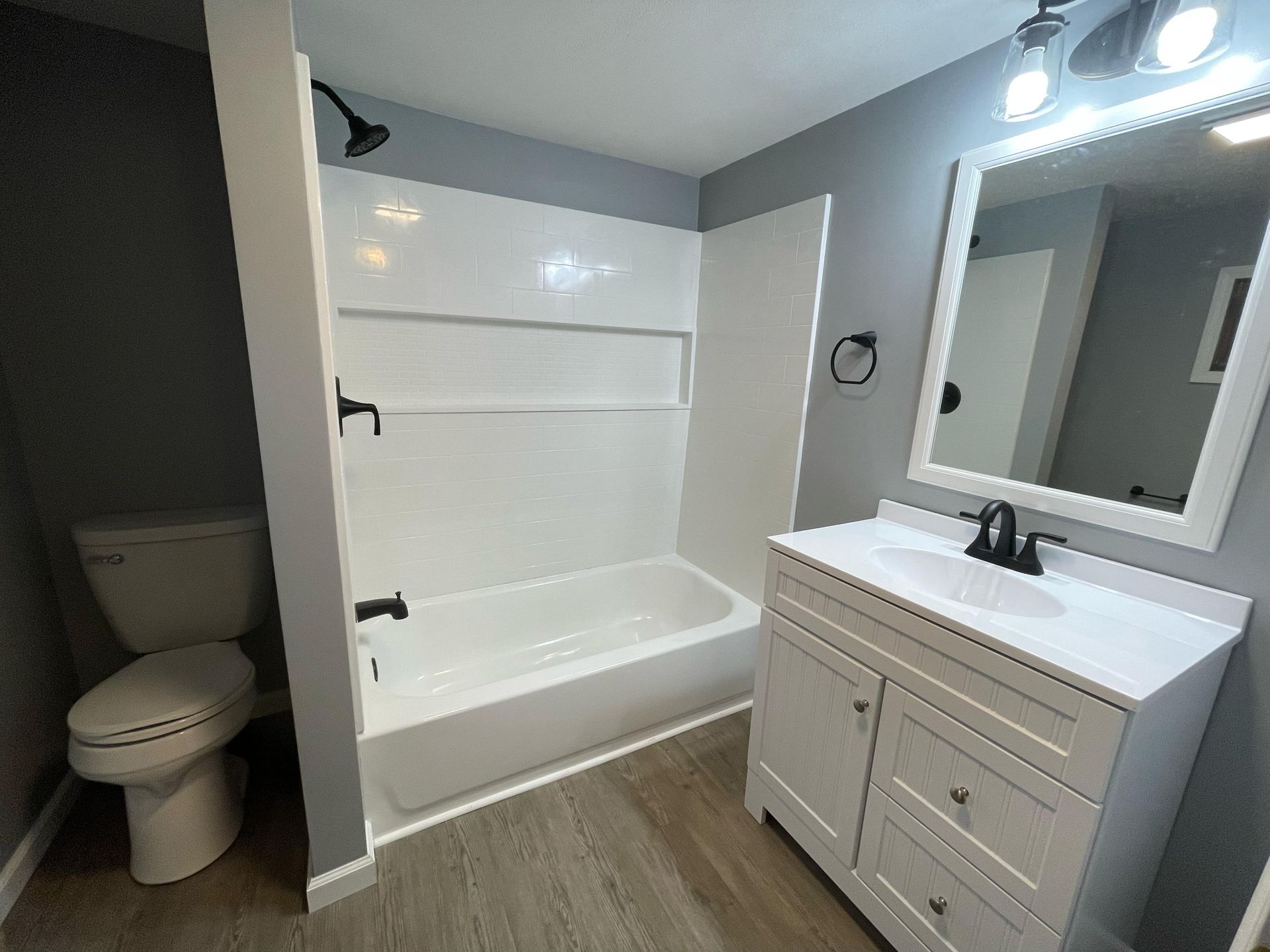Small bathroom with white tub-shower, toilet, and gray vanity with mirror and lights.
