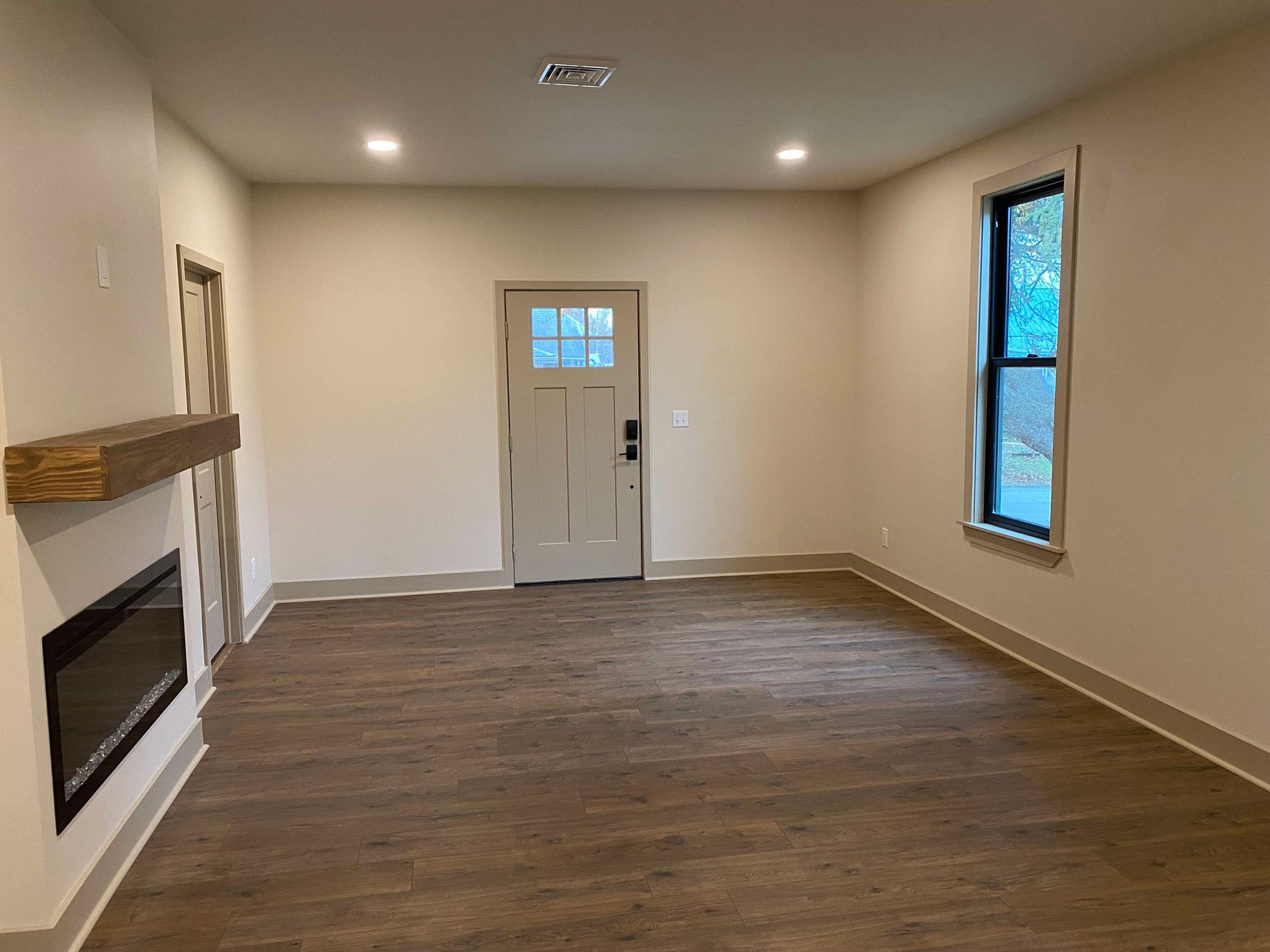 Empty living room with brown wood flooring, white walls, fireplace, front door, and a window.